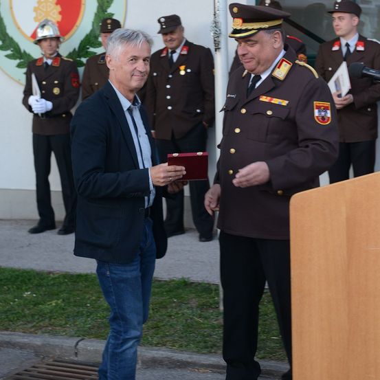 Ein Mann in einem Anzug erhält von einem Mann in Uniform eine rote Box, mit mehreren Militärangehörigen im Hintergrund.