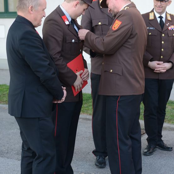 Eine formelle Zeremonie mit Militärangehörigen in braunen Uniformen; ein Offizier befestigt einen Abzeichen an einem jungen Mann, während andere zusehen.