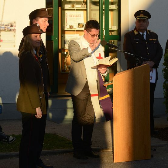 Ein Mann in Uniform liest von einem Buch an einem Podium, begleitet von zwei anderen in Uniform. Dahinter steht ein Gebäude mit Trophäen und einem Mikrofon.