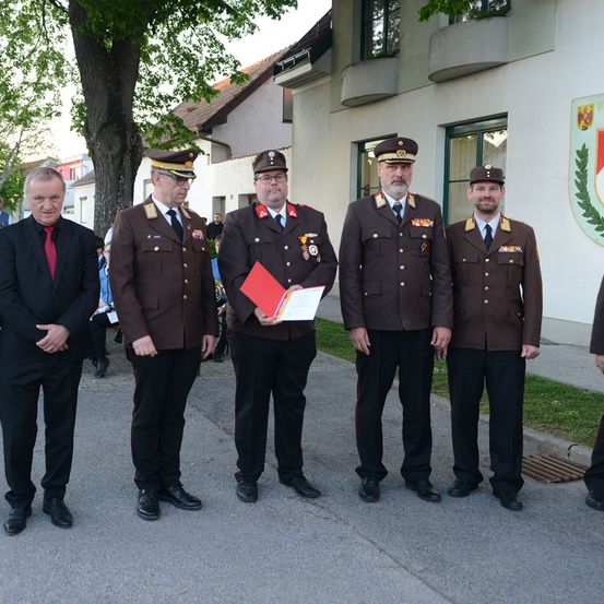 Fünf Männer in Uniform stehen vor einem Gebäude. Einer hält eine rote Mappe. Dahinter ein Gebäude mit Balkon und Baum.