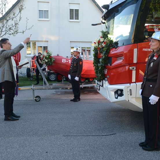Ein Feuerwehrmann in Uniform steht neben einem mit einem Kranz geschmückten Feuerwehrwagen. Ein Mann in Anzug hält ein Buch. Der Wagen ist mit B-1 beschriftet.