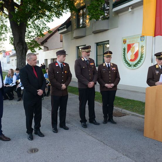 Fünf Männer in Uniform, einer in einem Anzug, stehen vor einem Gebäude mit einer Flagge und einem Logo. Dahinter sitzen mehrere Personen.