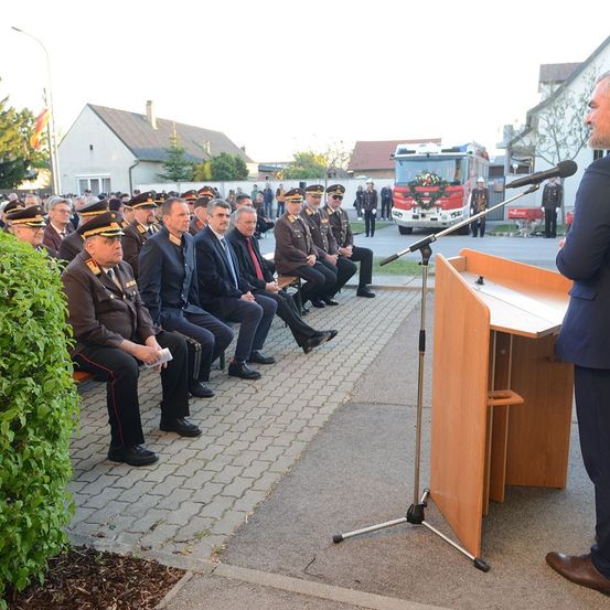 Ein Mann in Anzug hält eine Rede an einem Podium vor einer Gruppe uniformierter Beamter. Das Publikum sitzt auf Bänken und ist aufmerksam. Ein Feuerwehrauto ist hinter ihnen geparkt.