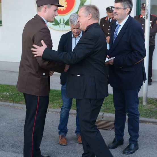 Drei Männer in Anzügen und einer in Uniform stehen draußen. Sie lächeln und schütteln Hände. Hinter ihnen ist eine Wand mit einem Logo und einige Leute stehen.