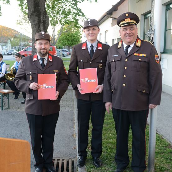 Drei Männer in Uniform, zwei halten rote Broschüren, stehen draußen. Dahinter sind ein Baum, ein Gebäude und Menschen zu sehen. Ein Mann spielt Trompete.