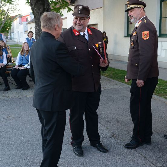 Ein Mann in einem Anzug übergibt ein Dokument einem uniformierten Beamten, der neben einem weiteren uniformierten Beamten vor einem Gebäude steht. Hinter ihnen sitzen Menschen auf einer Rasenfläche.