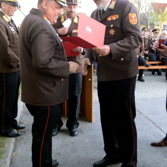 Zwei Männer in Militäruniformen tauschen bei einer Zeremonie im Freien eine rote Schachtel aus. Hinter ihnen stehen mehrere andere Offiziere in Uniform.