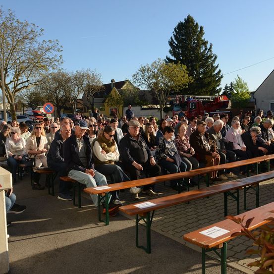 Eine Gruppe von Menschen sitzt auf Bänken unter Bäumen in einem Außenbereich, möglicherweise bei einer Veranstaltung. Im Hintergrund befinden sich Häuser und ein Feuerwehrfahrzeug.