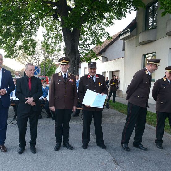 Fünf Männer in Uniform, einer mit einem Zertifikat, stehen vor einem Gebäude mit einem Baum. Dahinter sitzen zwei Personen an einem Tisch.