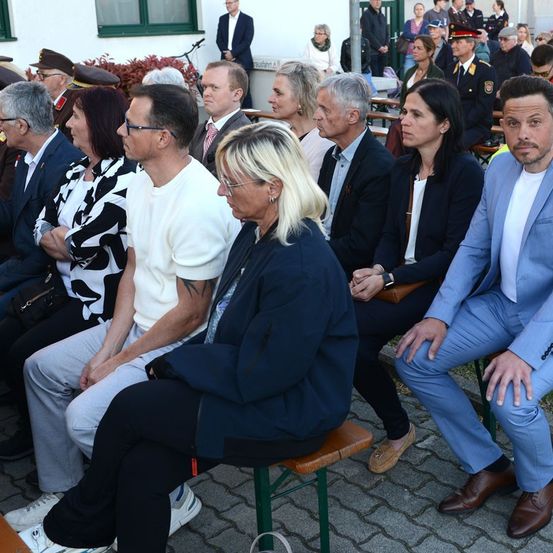 Eine Gruppe von Menschen in Anzügen und Kleidern sitzt auf Bänken. Eine Frau trägt eine Brille, eine andere hat ein Tattoo. Sie sind bei einer Veranstaltung im Freien.