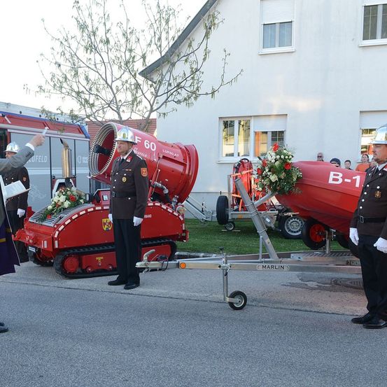 Eine Feuerwehrzeremonie mit uniformierten Beamten neben einem roten Fahrzeuganhänger mit Blumenschmuck, ein Gebäude mit Fenstern im Hintergrund.