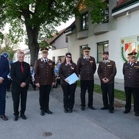 Eine Gruppe von Militärangehörigen und Beamten steht vor einem Gebäude, einer hält eine Mappe. Dahinter schauen Menschen zu, und ein Baum steht.