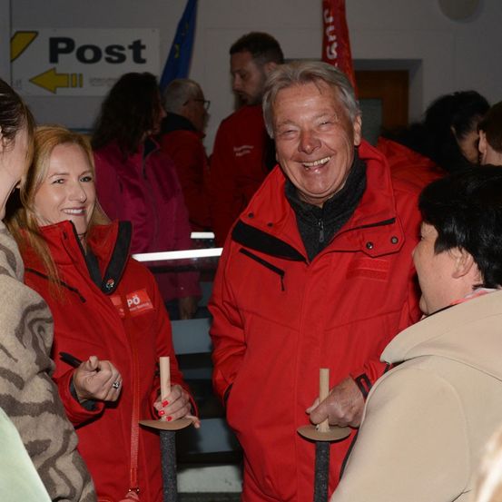 Eine Gruppe von Menschen in roten Jacken, darunter ein lächelnder Mann und eine Frau, versammelt sich in einem Raum, möglicherweise zu einer Feier.