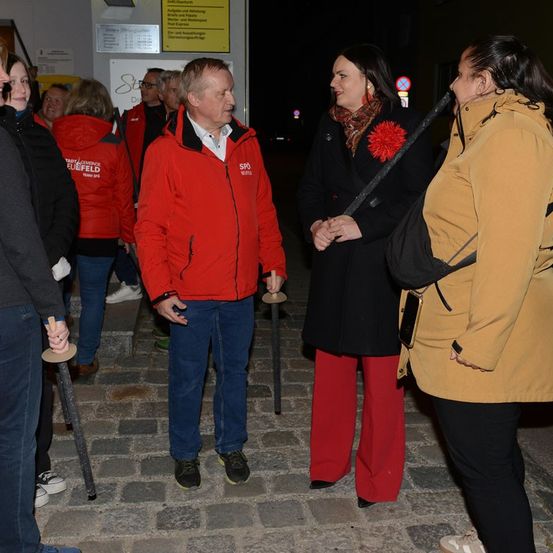 Eine Gruppe von Menschen in Winterkleidung steht bei Nacht draußen. Ein Mann in einem roten Mantel spricht mit einer Frau in einem schwarzen Mantel mit einer roten Blumenbrosche.
