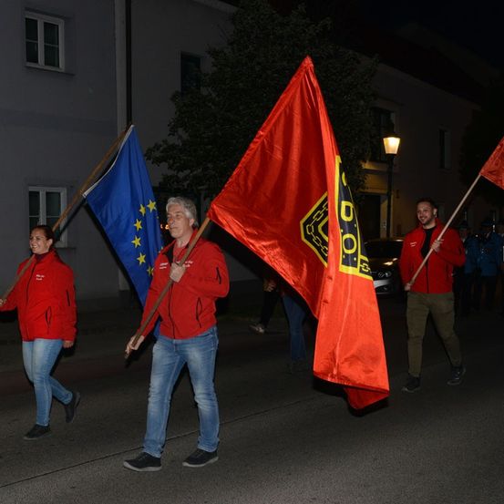 Drei Personen, zwei Männer und eine Frau, marschieren bei einer nächtlichen Parade. Sie tragen alle rote Jacken und halten Flaggen, eine davon ist die Flagge der Europäischen Union.