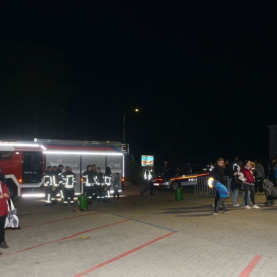 Eine Gruppe von Feuerwehrleuten steht bei Nacht vor einem Feuerwehrwagen. Mehrere Menschen sind versammelt, darunter ein Kind mit einer Taschenlampe. Ein Polizeiauto ist in der Nähe geparkt.