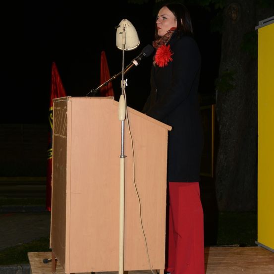 Eine Frau steht bei Nacht an einem Rednerpult mit Mikrofon, trägt einen schwarzen Mantel und rote Hosen und spricht. Hinter ihr sind ein Baum und Fahnen zu sehen.