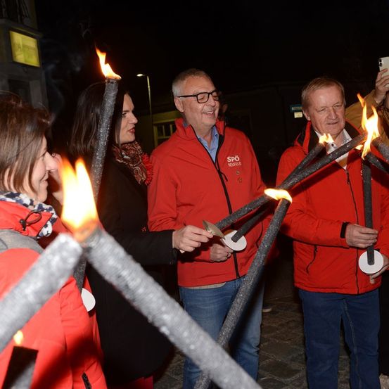Vier Personen in roten Jacken halten brennende Fackeln in einer nächtlichen Außenumgebung. Der Mann in der Mitte trägt eine Brille und ein Logo auf seiner Jacke. Die Frau links lächelt.