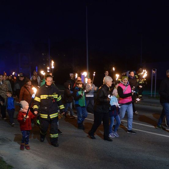 Eine Gruppe von Menschen, darunter Feuerwehrleute und Kinder, geht bei Nacht mit brennenden Fackeln in der Hand die Straße entlang. Der Hintergrund ist ein dunkler Himmel.