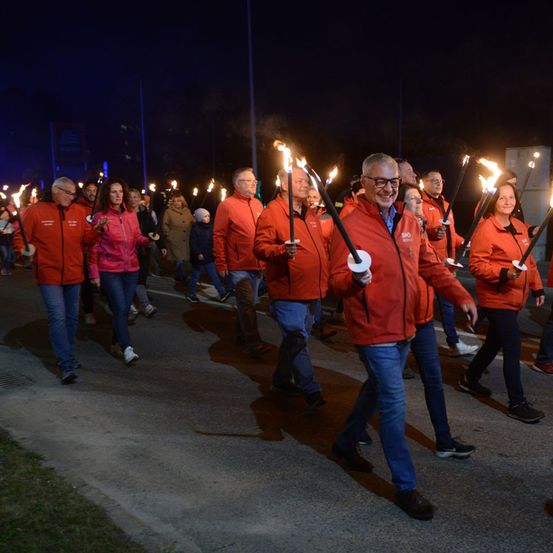 Eine Gruppe von Menschen in roten Jacken geht bei Nacht eine Straße entlang und hält brennende Fackeln.