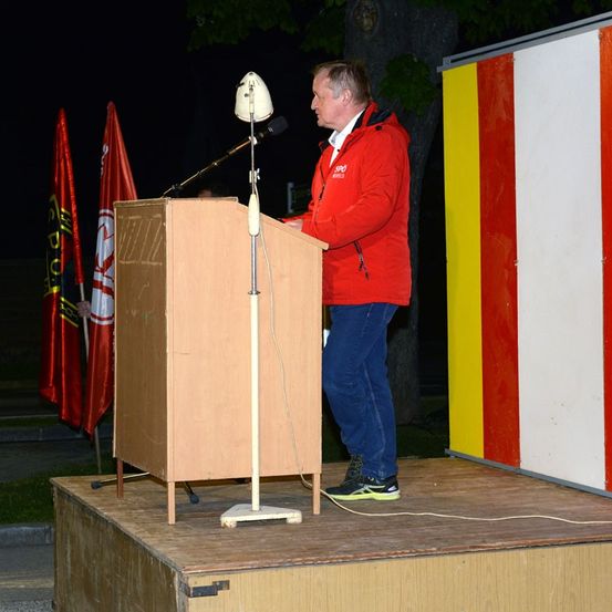 Ein Mann in einem roten Mantel steht bei Nacht an einem Rednerpult mit Mikrofon und hält wahrscheinlich eine Rede. Hinter ihm stehen zwei Fahnen und eine bunte Wand.