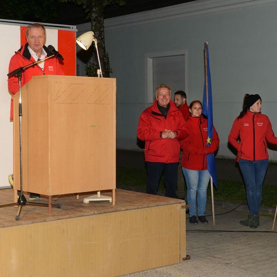Ein Mann steht an einem Rednerpult und spricht in ein Mikrofon, während drei Personen in roten Jacken hinter ihm stehen, eine hält eine Flagge. Die Szene spielt sich draußen bei Nacht ab.