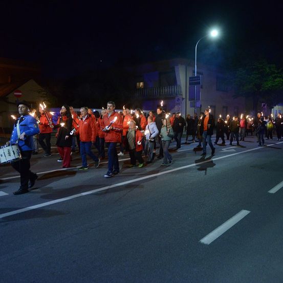 Eine Gruppe von Menschen in roten Kleidungsstücken marschiert bei Nacht die Straße hinunter und hält brennende Fackeln. Ein Trommler führt den Zug an.