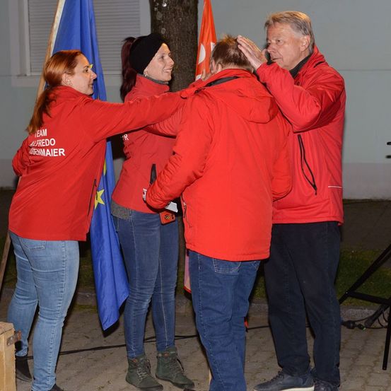 Drei Personen in roten Jacken stehen vor einer blauen Flagge und ein Mann legt seine Hand auf den Kopf einer Frau.