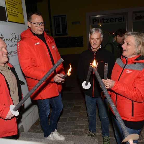 Vier Personen in roten Jacken halten brennende Fackeln in einer Außenumgebung, möglicherweise für eine zeremonielle Veranstaltung.