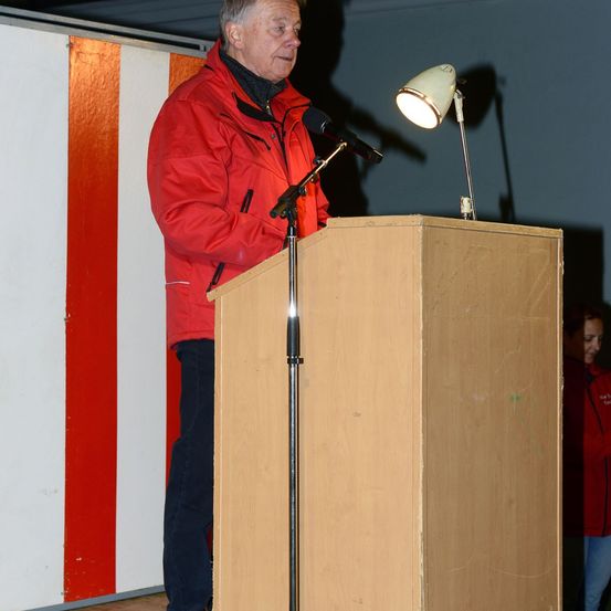 Ein älterer Mann in einer roten Jacke steht an einem Rednerpult mit Mikrofon und hält eine Rede. Eine Lampe steht auf dem Pult.