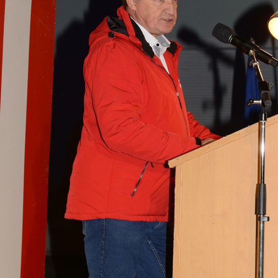 Ein Mann in einer roten Jacke steht an einem Rednerpult mit einem Mikrofon, möglicherweise hält er eine Rede.