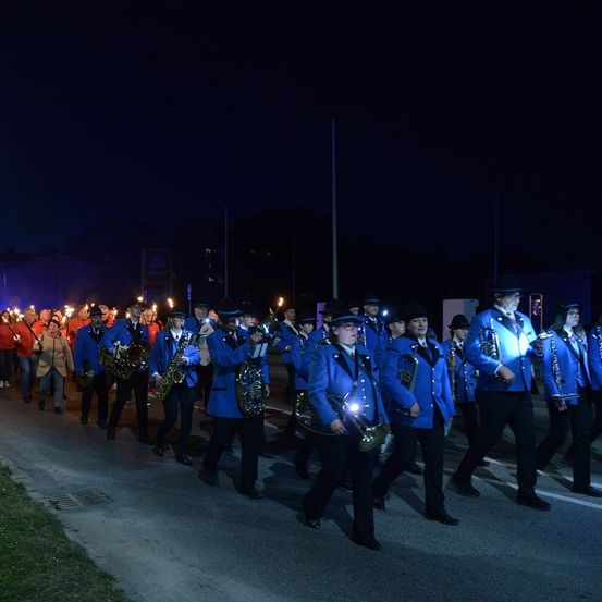 Eine Blaskapelle in blauen Uniformen und schwarzen Hüten spielt Instrumente und geht bei Nacht eine Straße entlang.