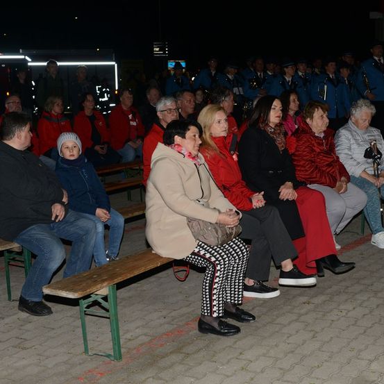 Eine Gruppe von Menschen sitzt nachts auf Bänken, einige tragen rote Jacken, möglicherweise bei einem Konzert oder einer Veranstaltung.