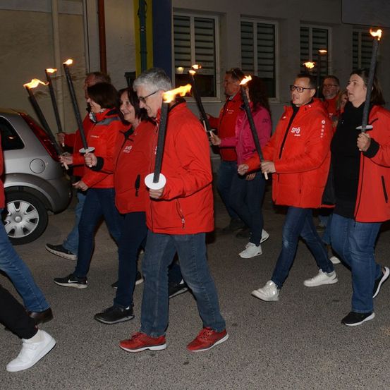 Eine Gruppe von Menschen in roten Jacken hält brennende Fackeln in der Hand und läuft nachts in einer Reihe auf der Straße.