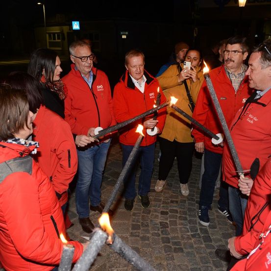 Eine Gruppe von Menschen in roten Jacken hält brennende Fackeln bei einer nächtlichen Veranstaltung im Freien.