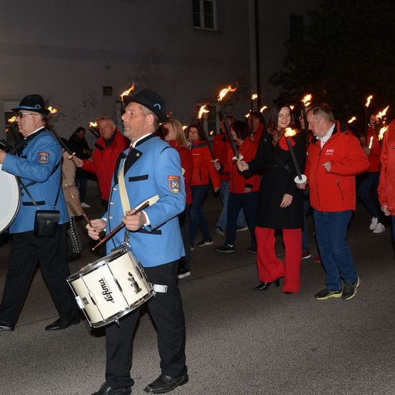 Eine Gruppe von Menschen in Blau und Rot gekleidet, die Fackeln halten und bei Nacht durch eine Parade ziehen.