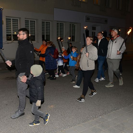 Eine Gruppe von Menschen zieht bei Nacht mit brennenden Fackeln in einem Umzug. Sie sind in Winterkleidung gekleidet und einige tragen Mützen. Die Straße ist nass und im Hintergrund sind Gebäude zu sehen.