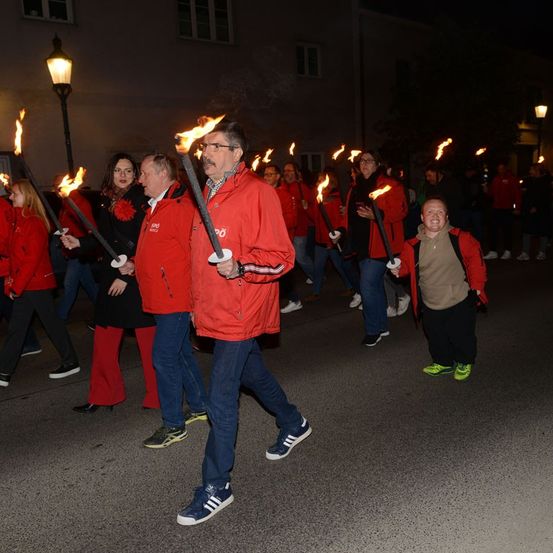 Eine Gruppe von Menschen in roten Jacken mit brennenden Fackeln geht nachts die Straße entlang.