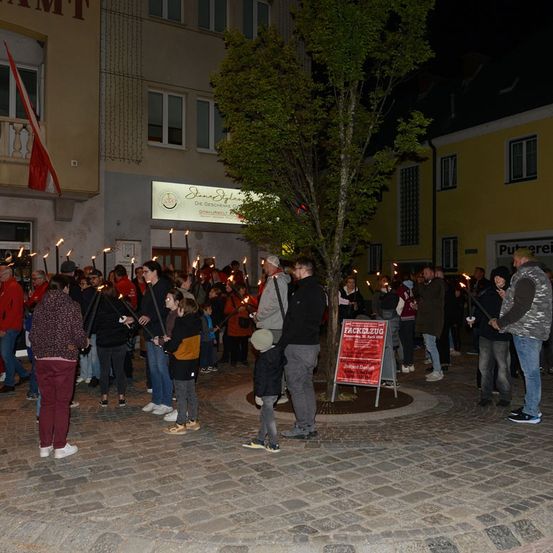 Eine Gruppe von Menschen mit brennenden Kerzen versammelt sich nachts auf einem Stadtplatz, mit Gebäuden und einem Baum im Hintergrund.