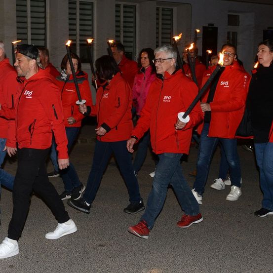 Eine Gruppe von Menschen in roten Jacken geht in einer Reihe und hält brennende Fackeln. Sie nehmen wahrscheinlich an einer Fackelprozession teil.