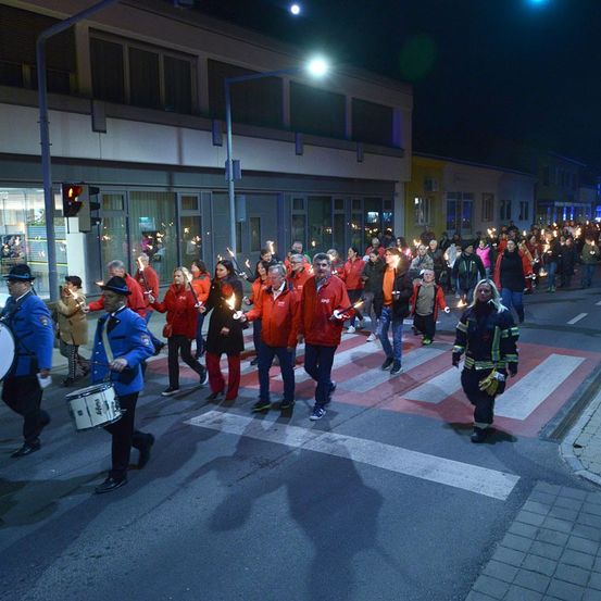 Ein nächtlicher Umzug mit Teilnehmern, die brennende Kerzen halten und rote Jacken tragen, marschiert über eine Straße, angeführt von einem Trommler.
