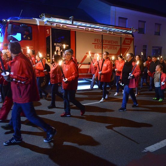 Eine Gruppe von Menschen in roten Jacken mit brennenden Fackeln in der Hand geht nachts in einer Reihe vor einem Feuerwehrwagen.
