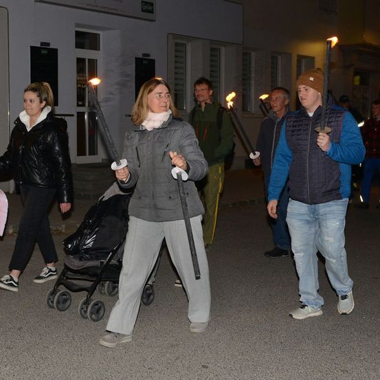 Eine Gruppe von Menschen geht nachts mit brennenden Fackeln in der Hand die Straße entlang. Eine Frau in einer grauen Jacke steht in der Mitte, während andere verschiedene Jacken und Turnschuhe tragen. Ein Kinderwagen steht hinter der Frau.