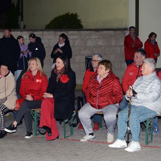 Eine Gruppe von Menschen in roten Jacken sitzt auf Bänken, einige stehen hinter einer Mauer, und sie alle lächeln.