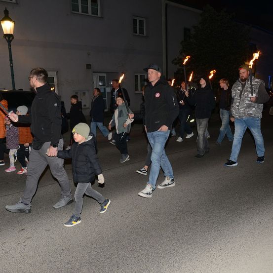 Eine Gruppe von Menschen, darunter Erwachsene und Kinder, zieht bei Nacht mit brennenden Fackeln in einer Prozession. Sie sind warm gekleidet, und die Szene spielt vor dem Hintergrund von Gebäuden und Bäumen.