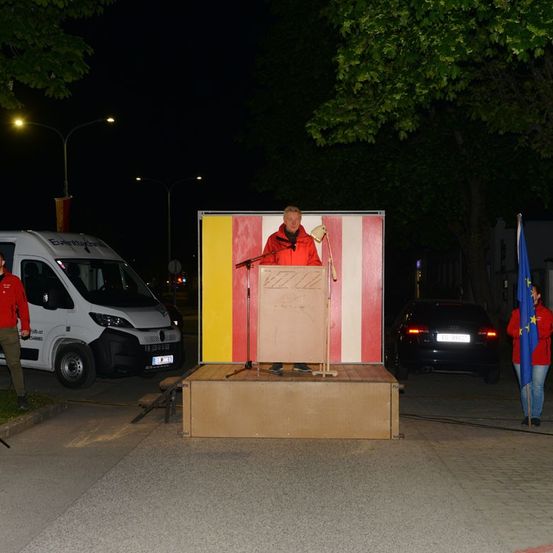 Ein Mann steht auf einer provisorischen Bühne an einem Rednerpult und spricht in ein Mikrofon. Ein weißer Lieferwagen und ein schwarzes Auto sind in der Nähe geparkt. Zwei Personen stehen rechts, eine hält eine Flagge. Bäume und Straßenlaternen sind im Hintergrund.