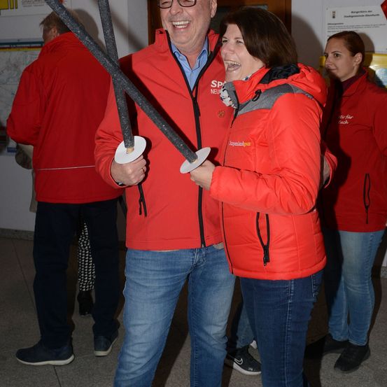 Ein Mann und eine Frau in roten Jacken halten Schwerter und lächeln, stehen in einem Flur mit anderen Leuten in roten Jacken.
