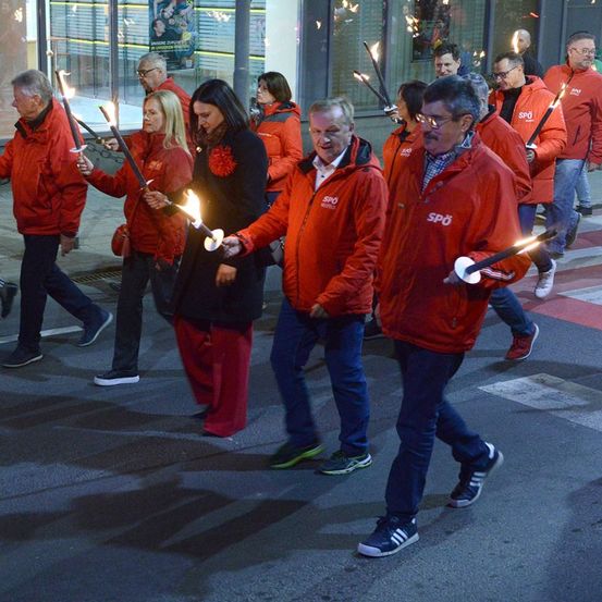 Eine Gruppe von Menschen in roten Jacken geht bei Nacht die Straße entlang und hält brennende Fackeln.