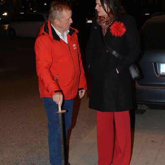 Ein Mann in einer roten Jacke und eine Frau in einem schwarzen Mantel mit einer roten Blumenbrosche stehen nachts auf einer Straße.