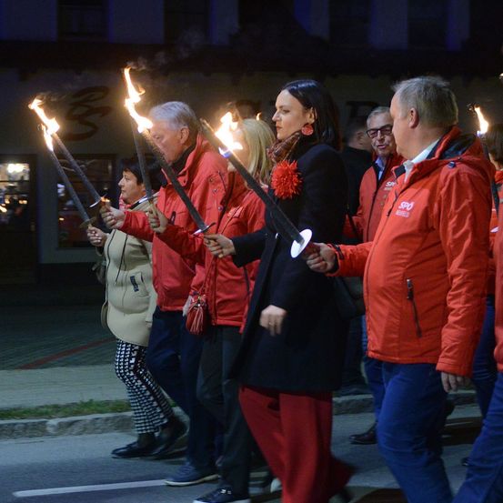 Eine Gruppe von Menschen in roten Jacken mit brennenden Fackeln geht bei Nacht die Straße entlang.